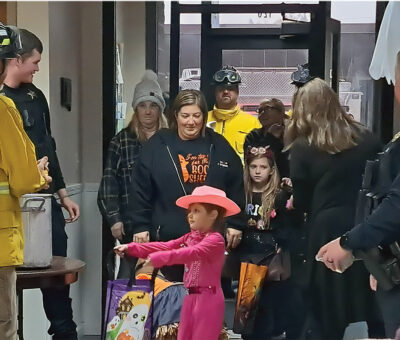 Young girl dressed as pink cowgirl holds out trick or treat bag to people dressed in firefighting gear